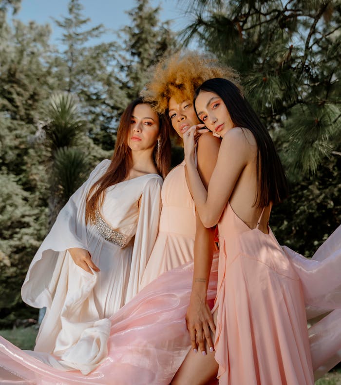 About Three women in elegant dresses pose outdoors, showcasing diverse fashion styles.